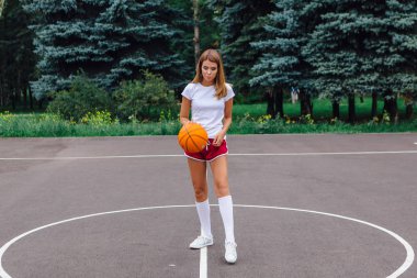 Güzel genç kız beyaz tişört, şort ve spor ayakkabısı, giyinmiş bir basketbol sahası üzerinde bir top ile çalış.