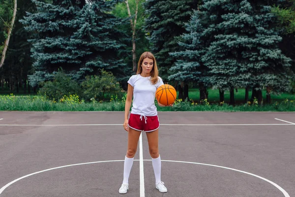 Güzel genç kız beyaz tişört, şort ve spor ayakkabısı, giyinmiş bir basketbol sahası üzerinde bir top ile çalış.