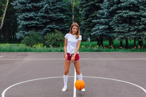 白の t シャツ、ショート パンツ、スニーカー、服を着た美しい少女はバスケット ボール コートでボールで遊ぶ. — ストック写真