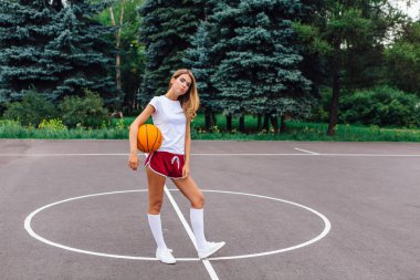 Güzel genç kız beyaz tişört, şort ve spor ayakkabısı, giyinmiş bir basketbol sahası üzerinde bir top ile çalış.