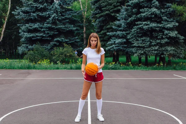 Mooi jong meisje gekleed in witte t-shirt, korte broek en sneakers, speelt met een bal op een basketbalveld. — Stockfoto