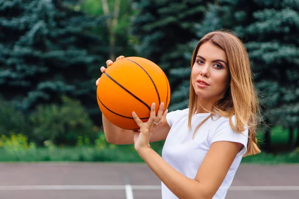 Gadis muda yang cantik mengenakan kaos putih, celana pendek dan sepatu olahraga, bermain dengan bola di lapangan basket . — Stok Foto