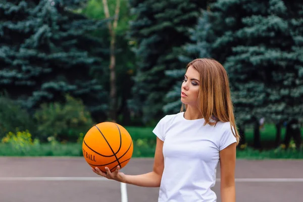 Gadis muda yang cantik mengenakan kaos putih, celana pendek dan sepatu olahraga, bermain dengan bola di lapangan basket . — Stok Foto