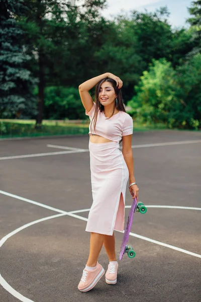 Retrato de una encantadora morena sonriente sosteniendo su monopatín en una cancha de baloncesto . — Foto de Stock