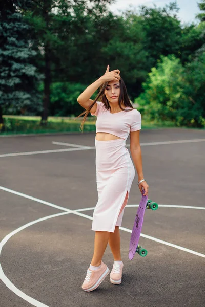 Retrato de una encantadora morena sonriente sosteniendo su monopatín en una cancha de baloncesto . — Foto de Stock