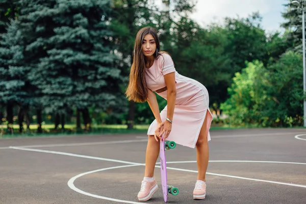 バスケット ボール コートで彼女のスケート ボードを持って笑顔の魅力的なブルネット女性の肖像画. — ストック写真