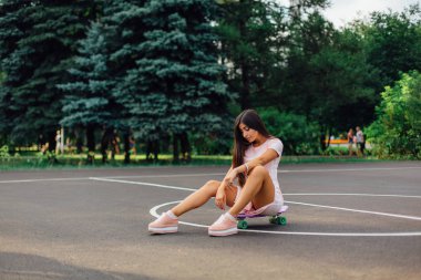 Basketbol sahasında kaykayında oturan gülümseyen çekici esmer bir kadının portresi. Modayı takip eden mutlu kadın gün batımında mola veriyor..