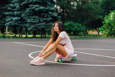 Basketbol sahasında kaykayında oturan gülümseyen çekici esmer bir kadının portresi. Modayı takip eden mutlu kadın gün batımında mola veriyor..