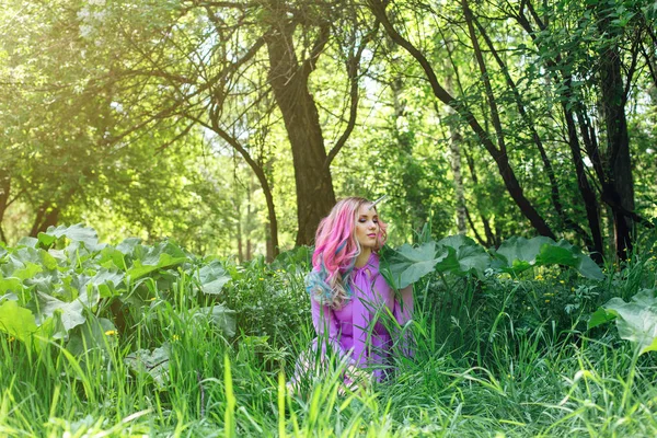 Fairy girl unicorn with rainbow hair and shiny makeup sitting on the grass in the forest.