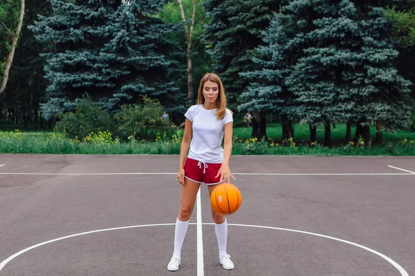 白いTシャツ ショートパンツ スニーカーを着た美しい若いブロンドの女の子は 屋外のバスケットボールコートでボールで遊んでいます スペースのコピー — ストック写真