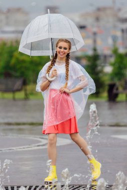Genç güzel kız sarı çizmeli iki örgüler ve şeffaf şemsiye ile Çeşme duruyor. Şehirde yağmurlu gün.