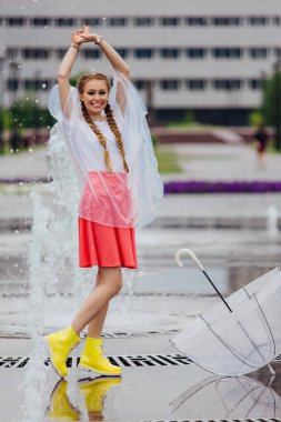 Genç güzel kız sarı çizmeli iki örgüler ve şeffaf şemsiye ile Çeşme duruyor. Şehirde yağmurlu gün.