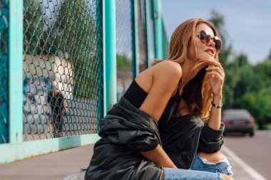 Moda portresi. Güneş gözlüğü takan genç bir kadın. Salonlu kot pantolon ve şehirde bombacı ceketi. Boşluğu kopyala.