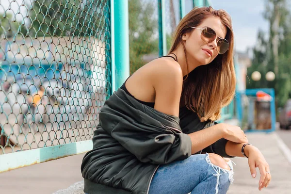 Retrato de moda de jovem na moda vestindo óculos de sol, jeans com salões e jaqueta de bombardeiro na cidade — Fotografia de Stock