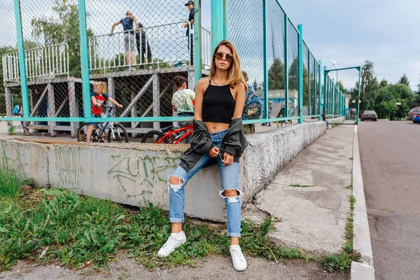 Retrato de moda de una joven de moda con gafas de sol, jeans con pasillos y chaqueta de bombardero en la ciudad —  Fotos de Stock