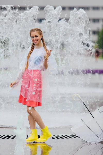 Joven chica bonita con dos trenzas en botas amarillas y con paraguas transparente se levanta cerca de la fuente . — Foto de Stock
