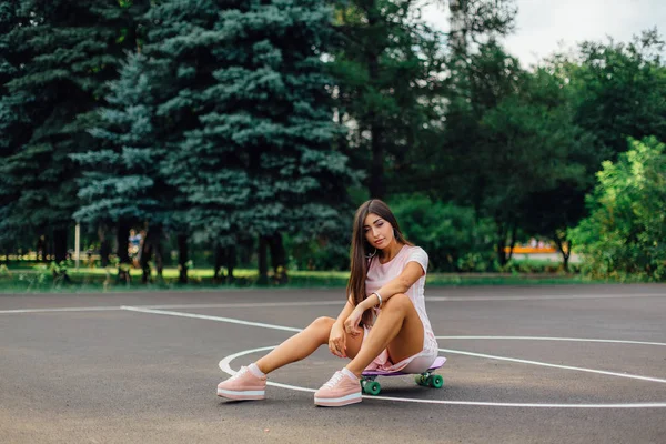 Portret van een lachende charmante brunette vrouw zit op haar skateboard op een basketbalveld. — Stockfoto