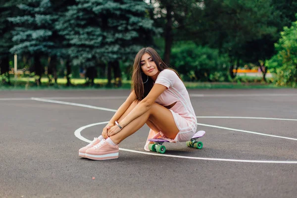 Portret van een lachende charmante brunette vrouw zit op haar skateboard op een basketbalveld. — Stockfoto