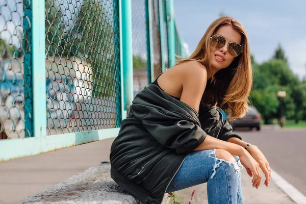 Retrato de moda de una joven de moda con gafas de sol, jeans con pasillos y chaqueta de bombardero en la ciudad —  Fotos de Stock