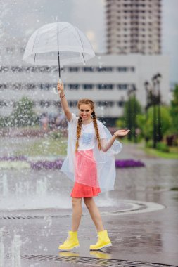 Genç güzel kız sarı çizmeli iki örgüler ve şeffaf şemsiye ile Çeşme duruyor. Şehirde yağmurlu gün.