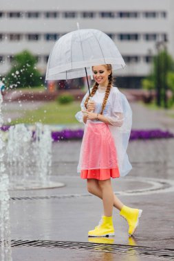 Genç güzel kız sarı çizmeli iki örgüler ve şeffaf şemsiye ile Çeşme duruyor. Şehirde yağmurlu gün.