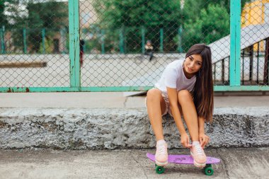 Yaz yaşam tarzı görüntü ile onun mor plastik kaykay kaykay coart yanında oturan ve ayakkabı bağcığı bağlama trendy genç ve güzel kız.