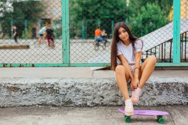 Yaz yaşam tarzı görüntü ile onun mor plastik kaykay kaykay coart yanında oturan ve ayakkabı bağcığı bağlama trendy genç ve güzel kız.