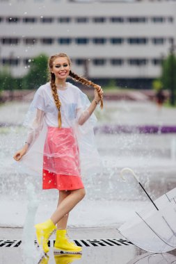 Genç güzel kız sarı çizmeli iki örgüler ve şeffaf şemsiye ile Çeşme duruyor. Şehirde yağmurlu gün.