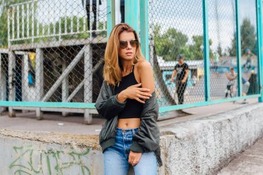 Moda portresi. Güneş gözlüğü takan genç bir kadın. Salonlu kot pantolon ve şehirde bombacı ceketi. Boşluğu kopyala.