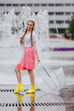 Genç güzel kız sarı çizmeli iki örgüler ve şeffaf şemsiye ile Çeşme duruyor. Şehirde yağmurlu gün.