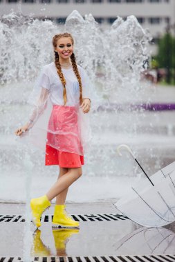 Genç güzel kız sarı çizmeli iki örgüler ve şeffaf şemsiye ile Çeşme duruyor. Şehirde yağmurlu gün.