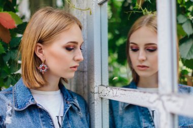 Moda kız waching yansıması, büyük boy kot ceket ve güzel kulak halkaları, ayna pencere ayakta.