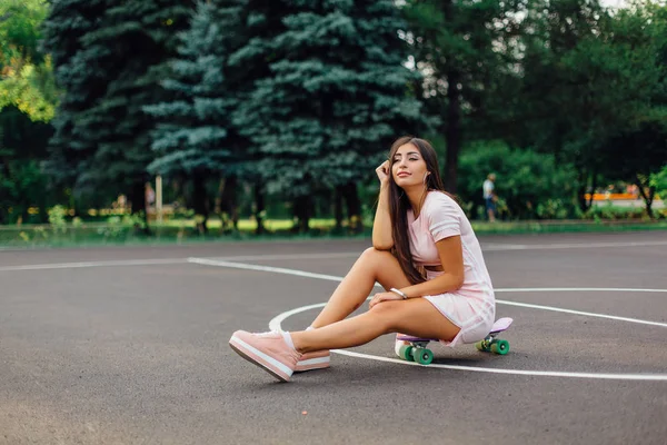 Porträtt av en leende charmiga brunett kvinna sitter på hennes skateboard på en basketplan. Stockfoto