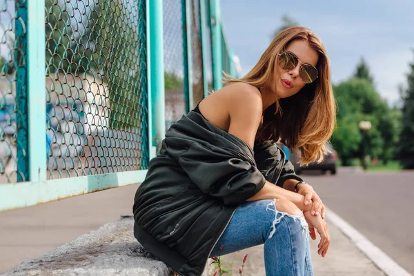 Portrait de mode de jeune femme branchée portant des lunettes de soleil, un jean avec des salles et une veste de bombardier dans la ville — Photo