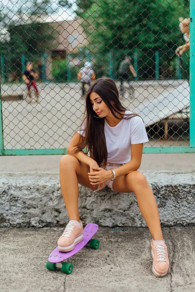 Summer lifestyle image of trendy pretty young girl sitting next to the skateboard coart with her plastic skateboard. — Stock Photo, Image
