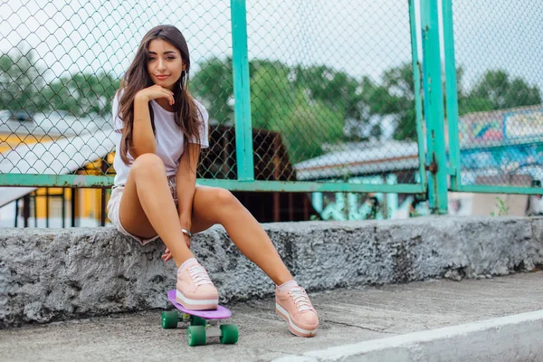 Zomer levensstijl foto van trendy mooie jonge meisje zit naast de skateboard coart met haar kunststof skateboard. — Stockfoto