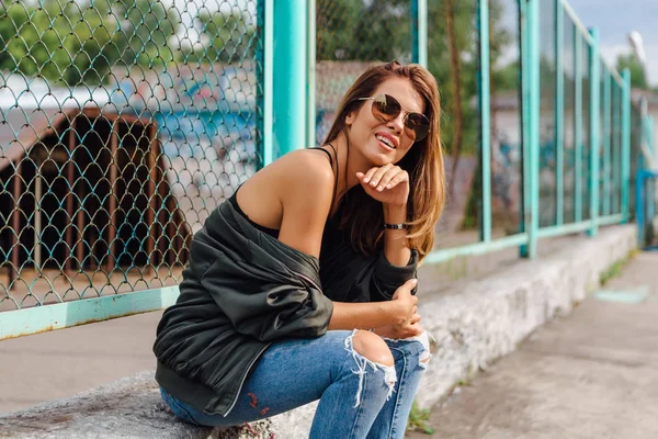 Fashion portrait of trendy young woman wearing sunglasses, jeans with halls and bomber jacket in the city — Stock Photo, Image