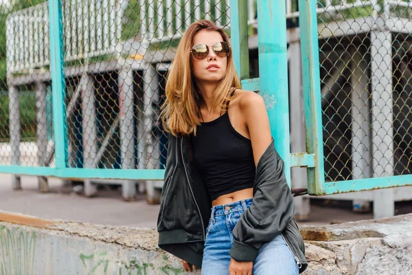 Mode portret van trendy jonge vrouw dragen van een zonnebril, jeans met zalen en blouson in de stad — Stockfoto