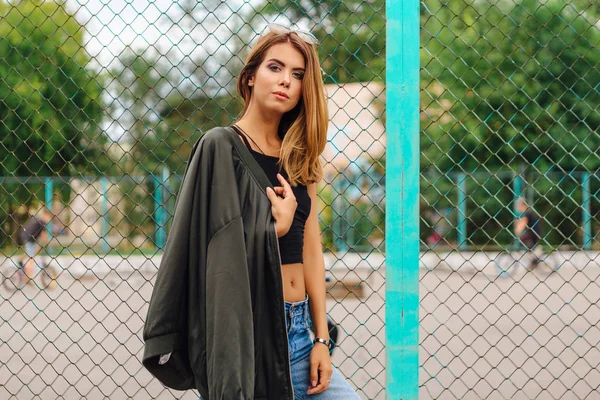 Retrato de moda de una joven de moda con gafas de sol, jeans con pasillos y chaqueta de bombardero en la ciudad —  Fotos de Stock