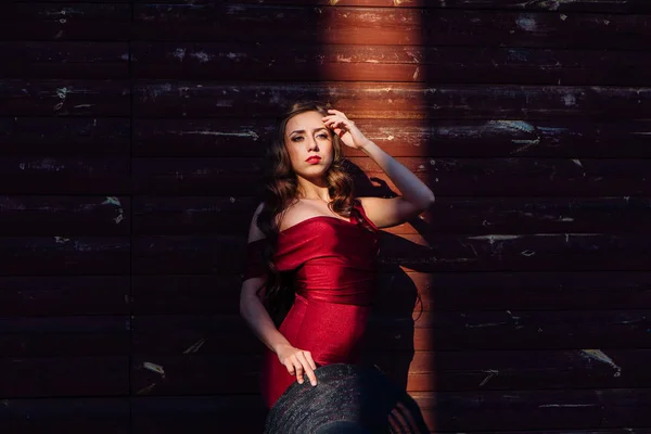Hermosa joven con un largo vestido de noche rojo con un tren y sombrero negro de pie en un rayo de luz — Foto de Stock