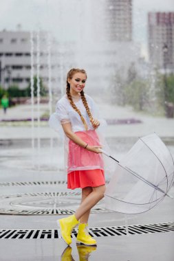 Genç güzel kız sarı çizmeli iki örgüler ve şeffaf şemsiye standları Çeşme yakınındaki.