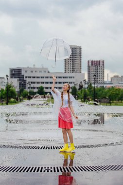 Genç güzel kız sarı çizmeli iki örgüler ve şeffaf şemsiye standları Çeşme yakınındaki.