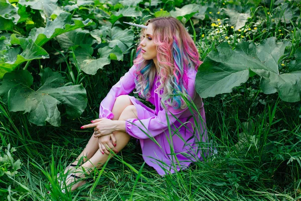 Fairy girl unicorn with rainbow hair and shiny makeup sitting on the grass.