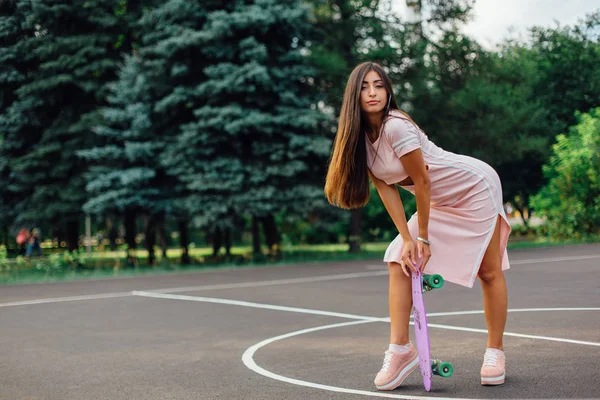 Retrato de uma morena encantadora sorridente segurando seu skate em um campo de basquete . — Fotografia de Stock