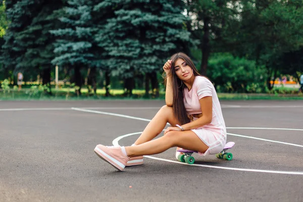 Portret van een lachende charmante brunette vrouw zit op haar skateboard op een basketbalveld. — Stockfoto