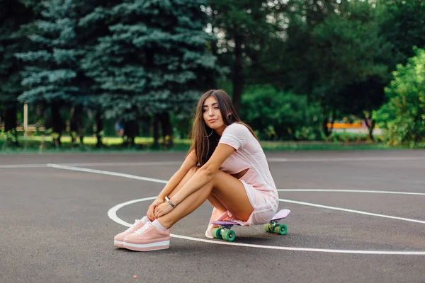 Retrato de uma morena encantadora sorridente sentada em seu skate em uma quadra de basquete . — Fotografia de Stock