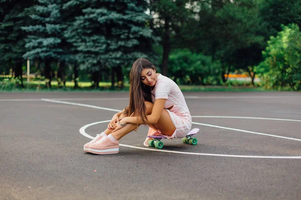 Portret van een lachende charmante brunette vrouw zit op haar skateboard op een basketbalveld. — Stockfoto