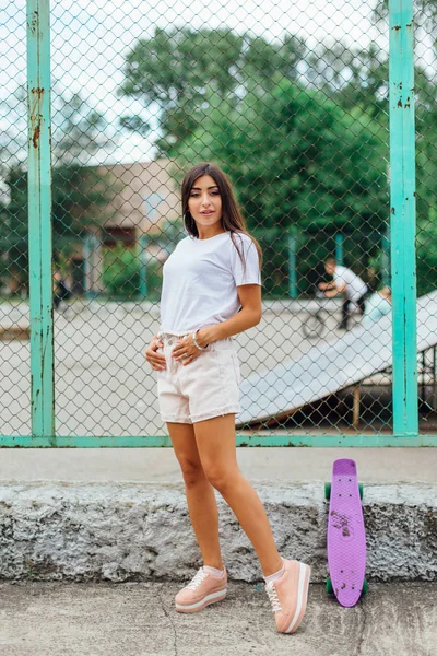 Summer lifestyle image of trendy pretty young girl standing next to the scateboard coart with her plastic scateboard. — Stock Photo, Image