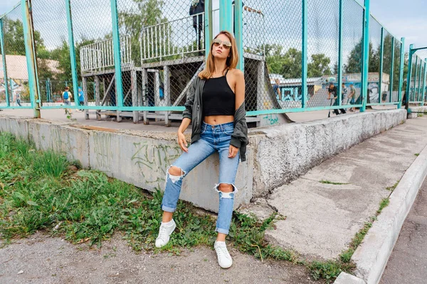 Potret busana wanita muda trendi mengenakan kacamata hitam, celana jeans dengan aula dan jaket pembom di kota — Stok Foto