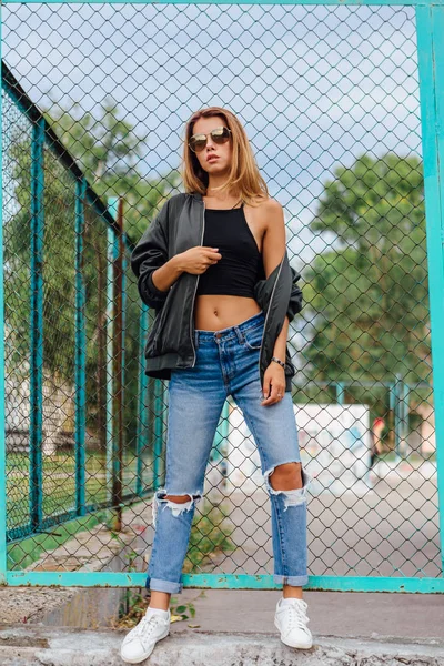Модный портрет модной молодой женщины в солнцезащитных очках, джинсах с залами и куртке подрывника в городе — стоковое фото